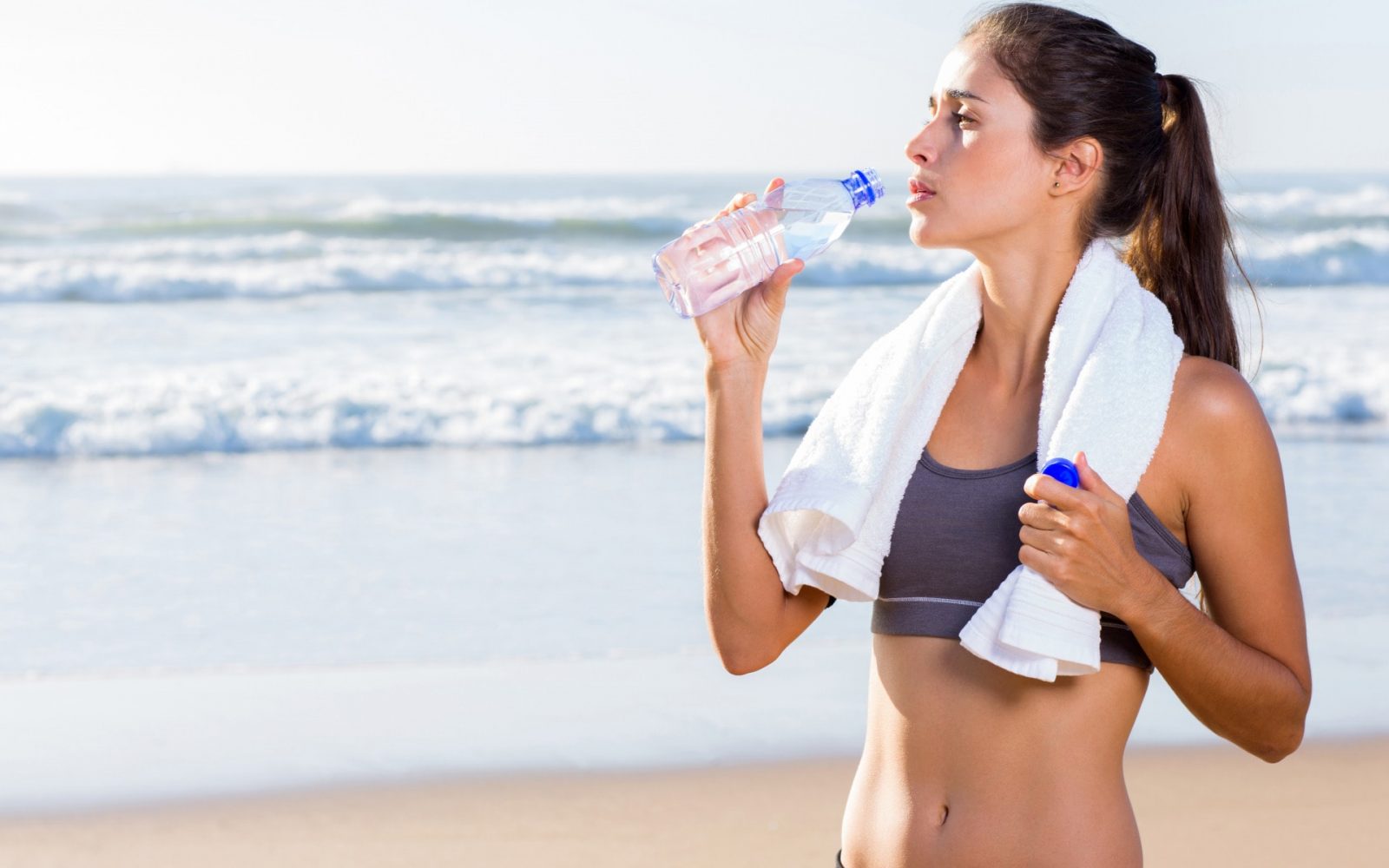 Девушка на берегу море с полотенцем на шеи и голым пузом пьет чистую воду из бутылке